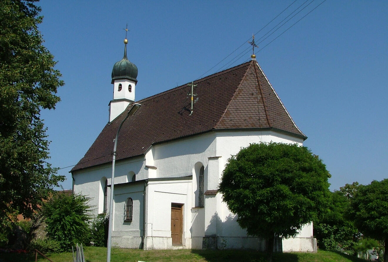 Weißes Kirchengebäude mit einem hohen, grünen Spitzdach und einem Zwiebelturm, umgeben von Bäumen.