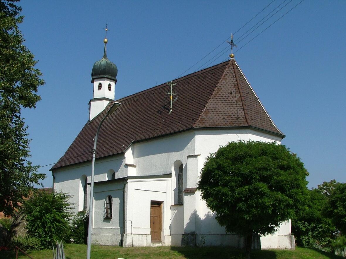 Weißes Kirchengebäude mit einem hohen, grünen Spitzdach und einem Zwiebelturm, umgeben von Bäumen.