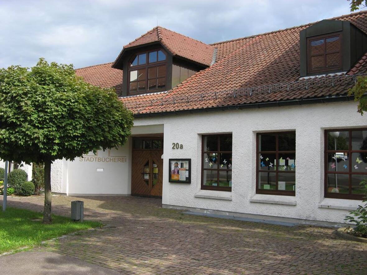 Eingang der Stadtbücherei mit großen Fenstern und einem Baum im Vordergrund.