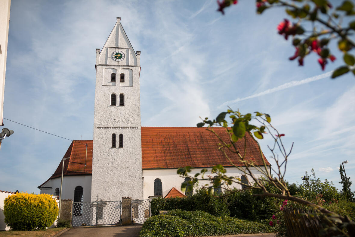 Weißer Kirchturm mit rotem Dach und Uhr, umgeben von Grünpflanzen und blühenden Büschen.