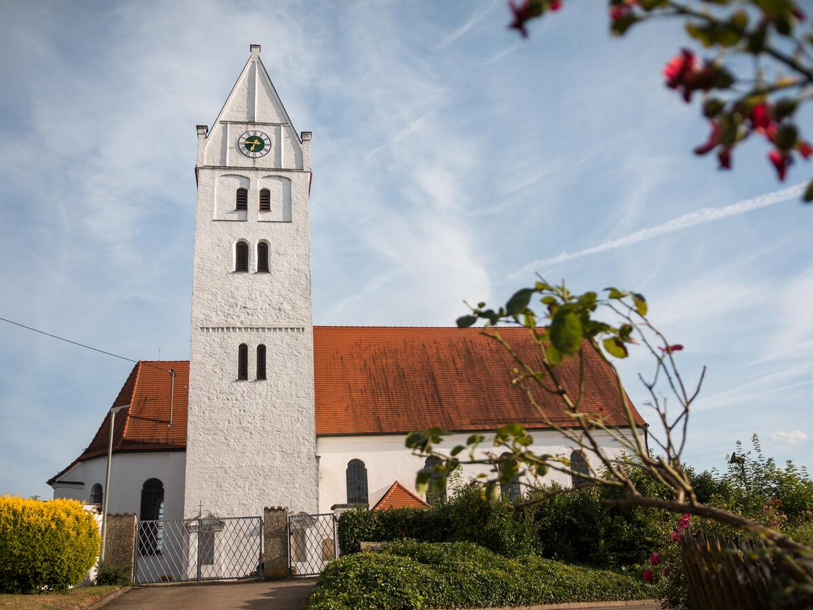 Weißer Kirchturm mit rotem Dach und Uhr, umgeben von Grünpflanzen und blühenden Büschen.