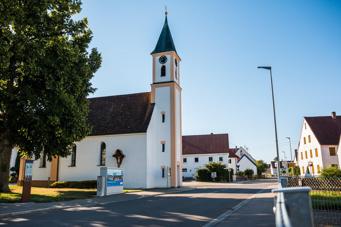 Kirche mit spitzem Turm und Ziffernblatt in einer ruhigen Straße, umgeben von Häusern.