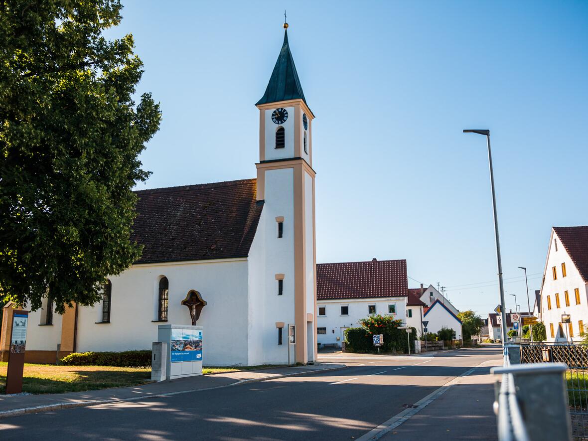 Kirche mit spitzem Turm und Ziffernblatt in einer ruhigen Straße, umgeben von Häusern.