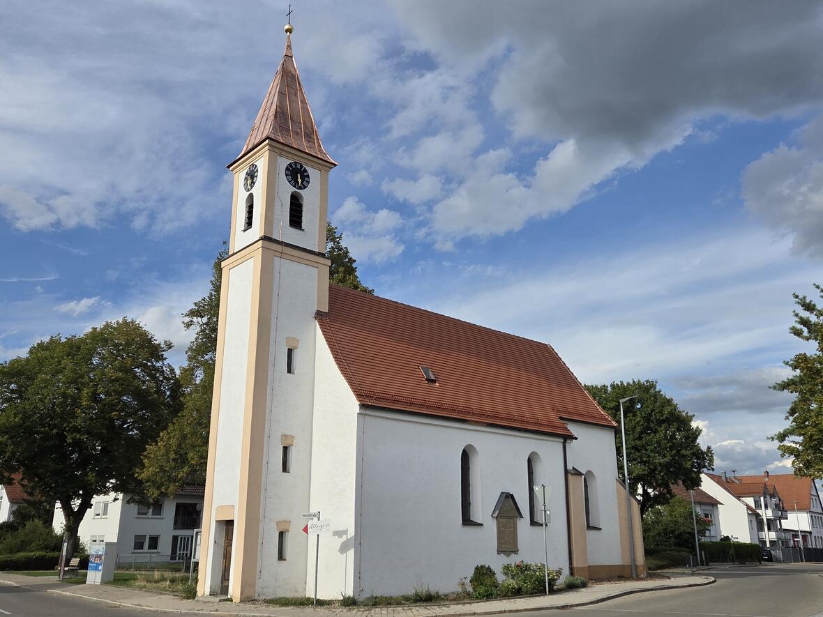 Kirche mit spitzem Turm und Ziffernblatt in einer ruhigen Straße, umgeben von Häusern.