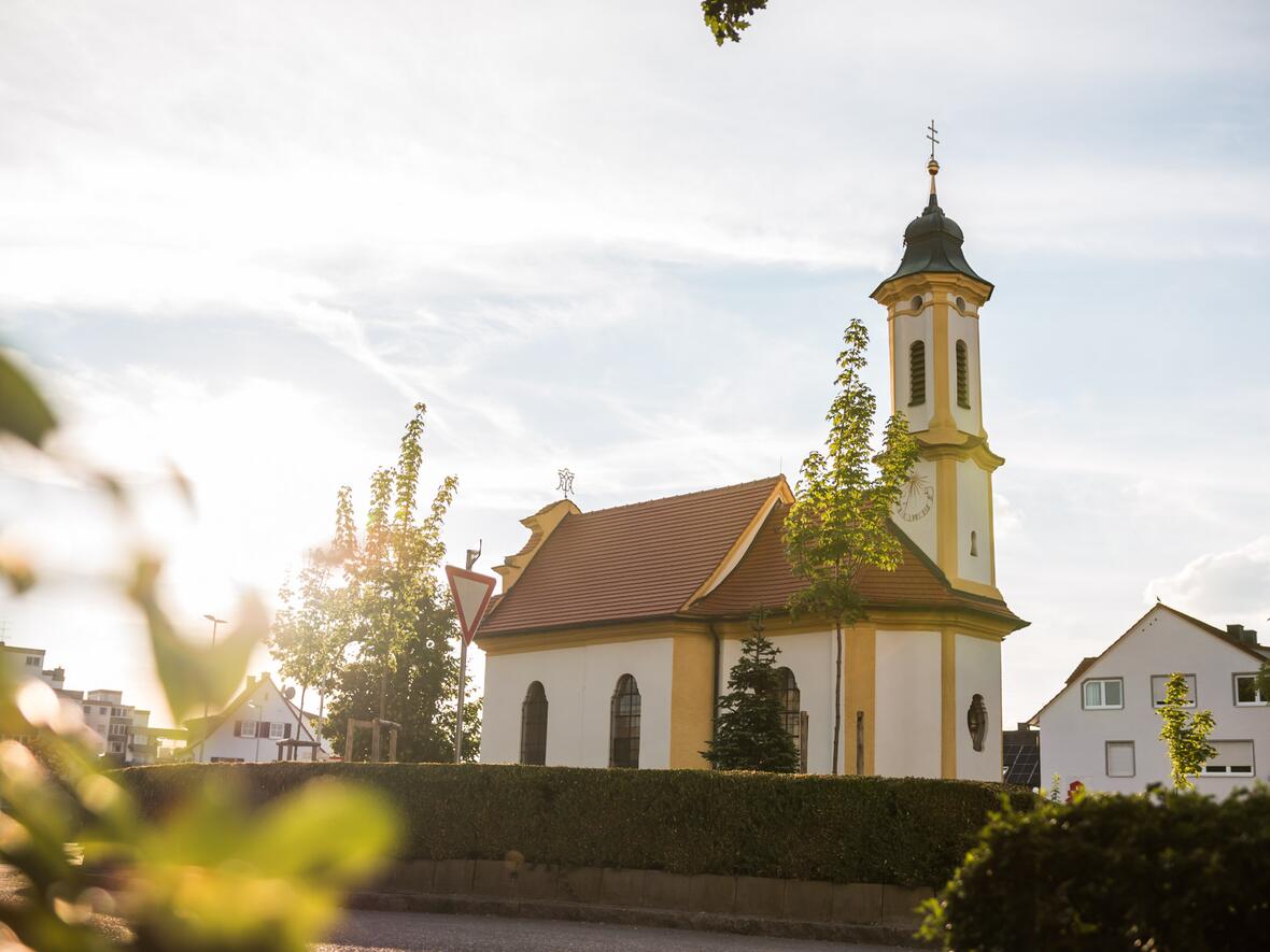 Die Kapelle im Morgenlicht, umgeben von Bäumen und einem gepflegten Grünstreifen.