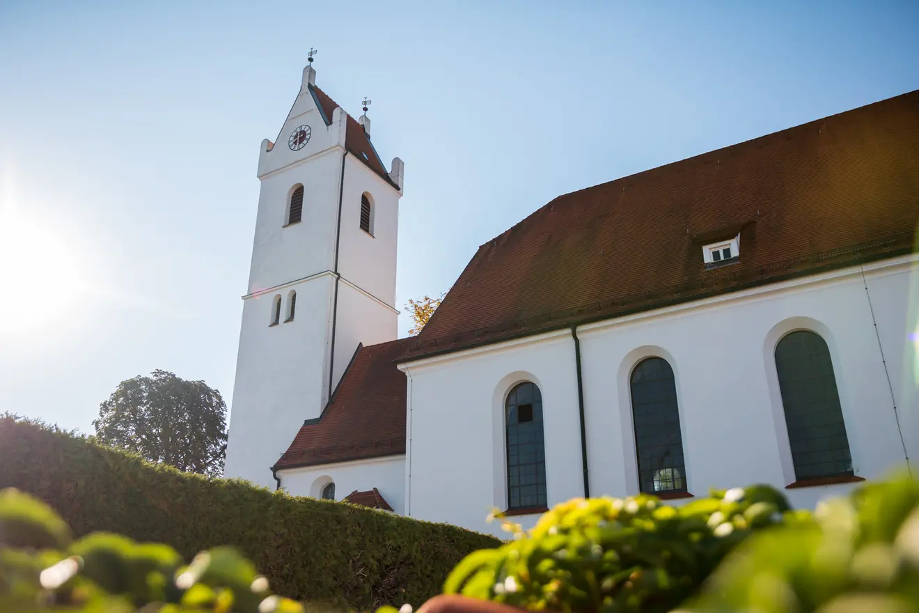 Kirche mit markantem Turm und weißen Wänden, umgeben von grüner Vegetation im Sonnenlicht.