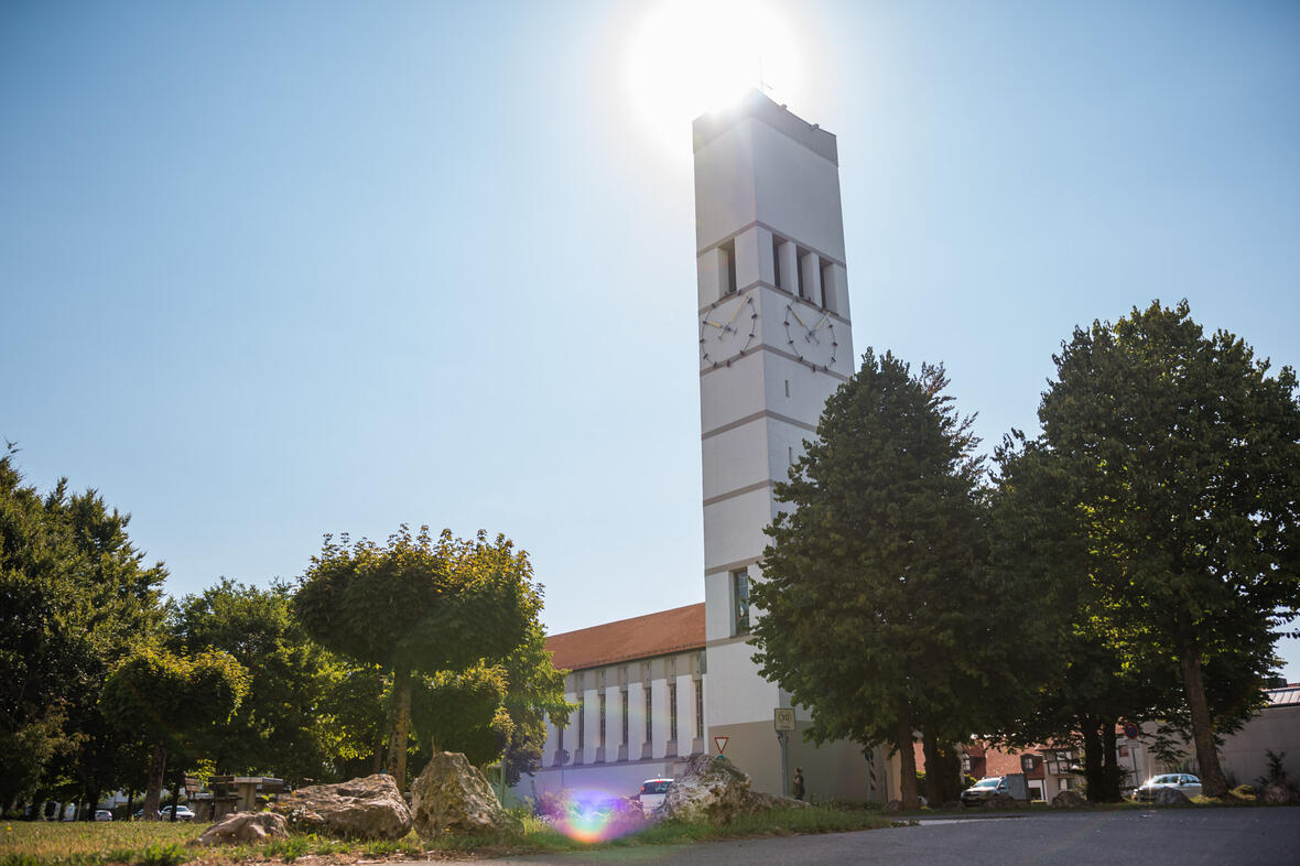 Kirchturm mit Uhr, bei Sonnenlicht, umgeben von Bäumen und einem klaren Himmel.