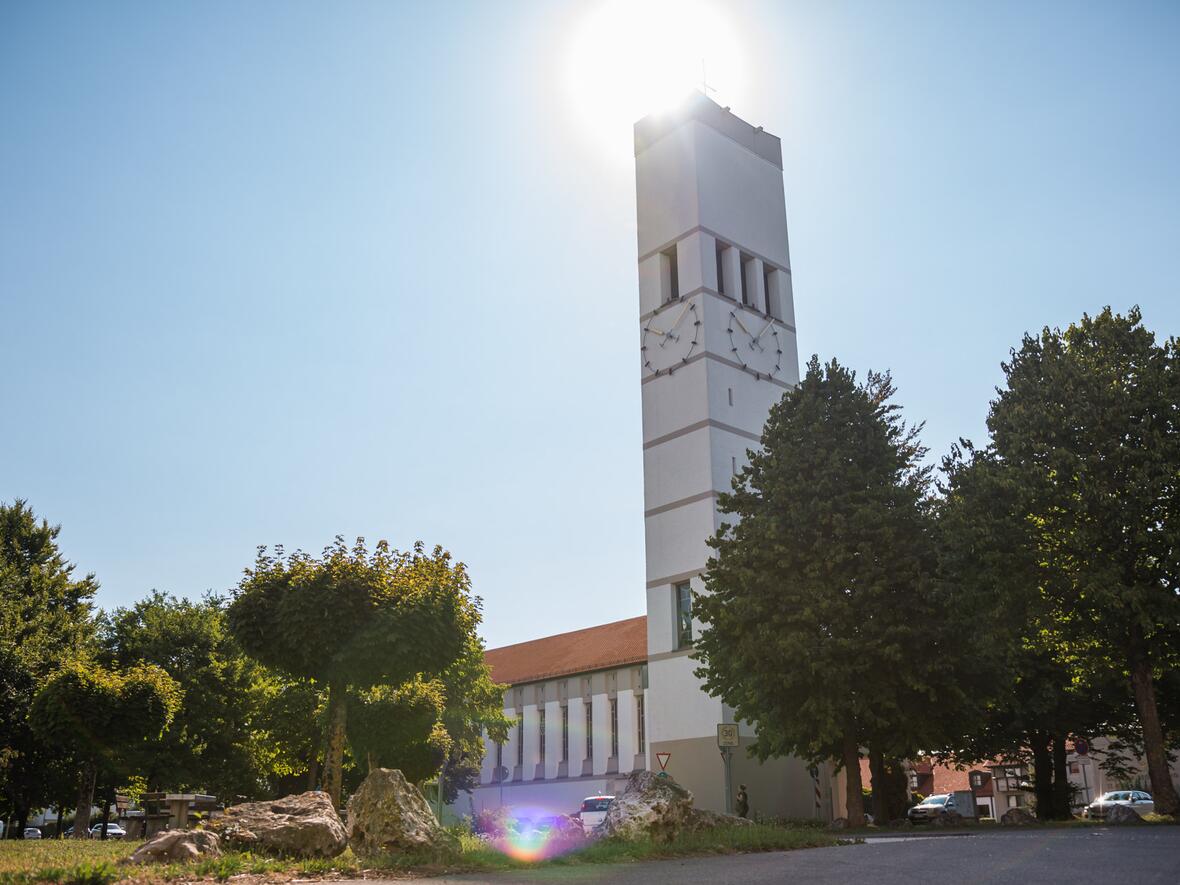 Kirchturm mit Uhr, bei Sonnenlicht, umgeben von Bäumen und einem klaren Himmel.