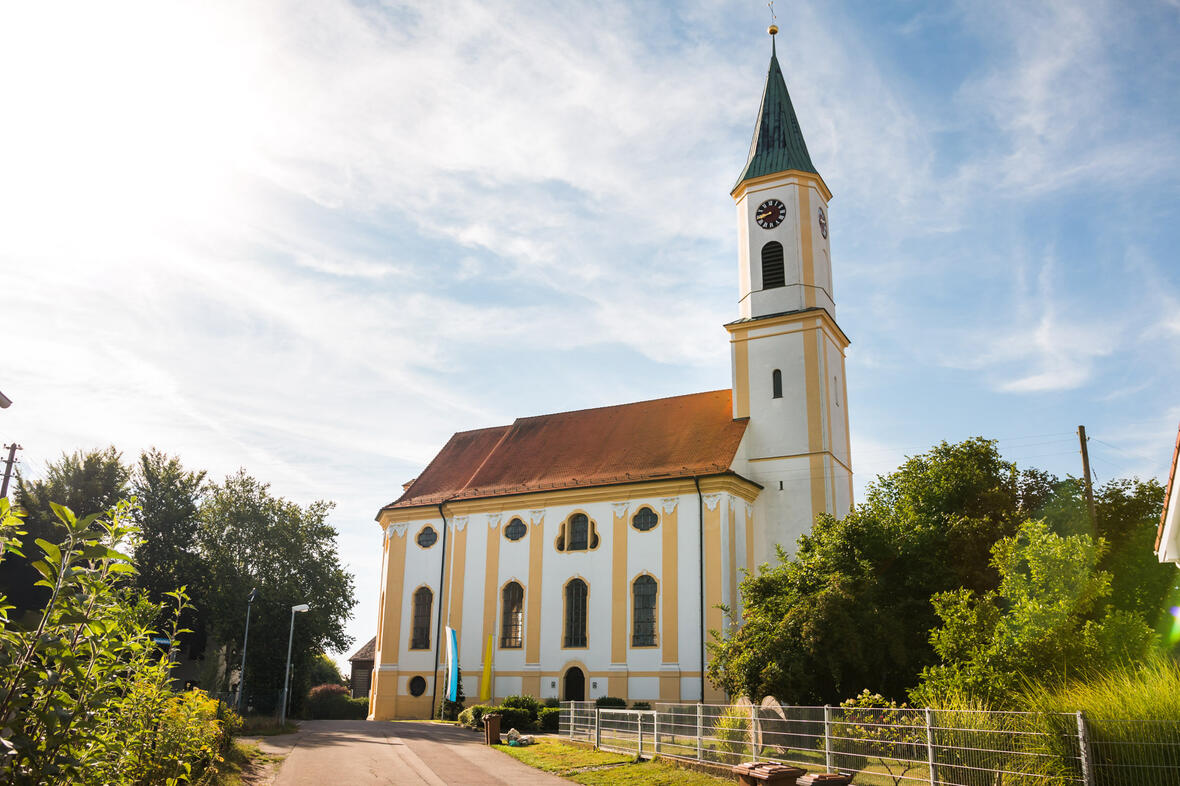 Kirche in ruhiger Umgebung, im Sonnenlicht, umgeben von Bäumen und einem Feldweg.